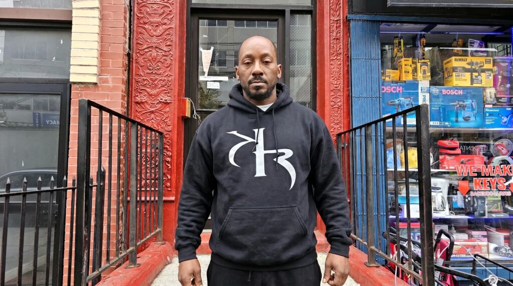 Commander KR standing outside a Bronx storefront in a KR Worldwide monogram hoodie.